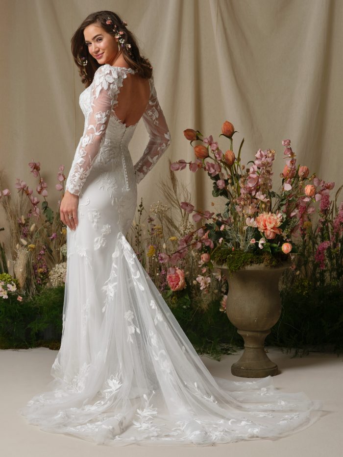 Aubra by Rebecca Ingram lace sheath wedding dress featuring an illusion open back, floral lace detailing, long sheer sleeves, covered buttons, and a flowing tulle train.