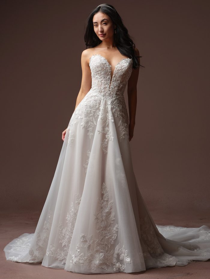 Front view of a model wearing the Bernadine A-line Wedding Dress by Rebecca Ingram in ivory, featuring a strapless plunging sweetheart neckline with floral lace appliqués, a fitted lace bodice, and a flowing tulle A-line skirt scattered with lace floral motifs and a train. 