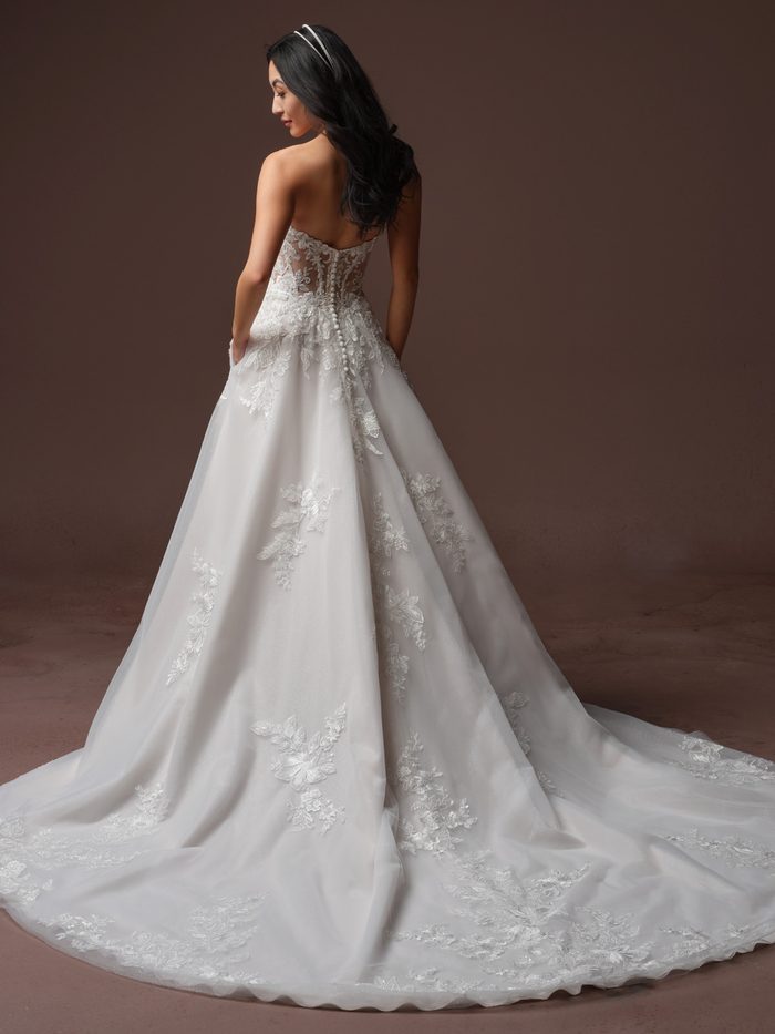 Back view of a model wearing the Bernadine A-line Wedding Dress by Rebecca Ingram in ivory, showing an illusion lace back with a row of covered buttons down the spine, and a flowing tulle A-line skirt with scattered lace floral appliqués and a long train.