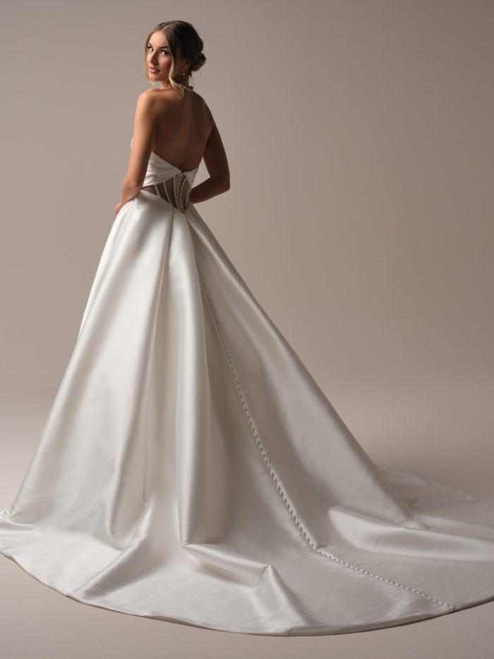 Back view of a bride wearing the Brooke ballgown wedding dress by Rebecca Ingram, showing a strapless satin bodice with a low back, corset-style side cutouts, and a full satin skirt with a covered-button trim on the train.