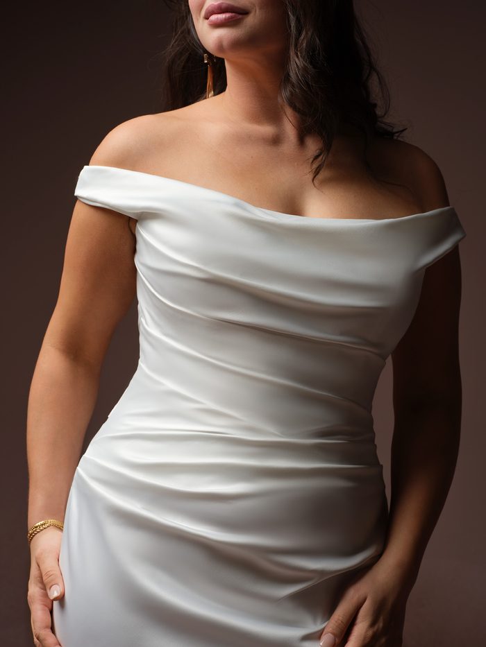 Close-up of the Clementine sheath wedding dress by Rebecca Ingram, showing the draped off-the-shoulder satin neckline and ruched bodice detail on a curve model.