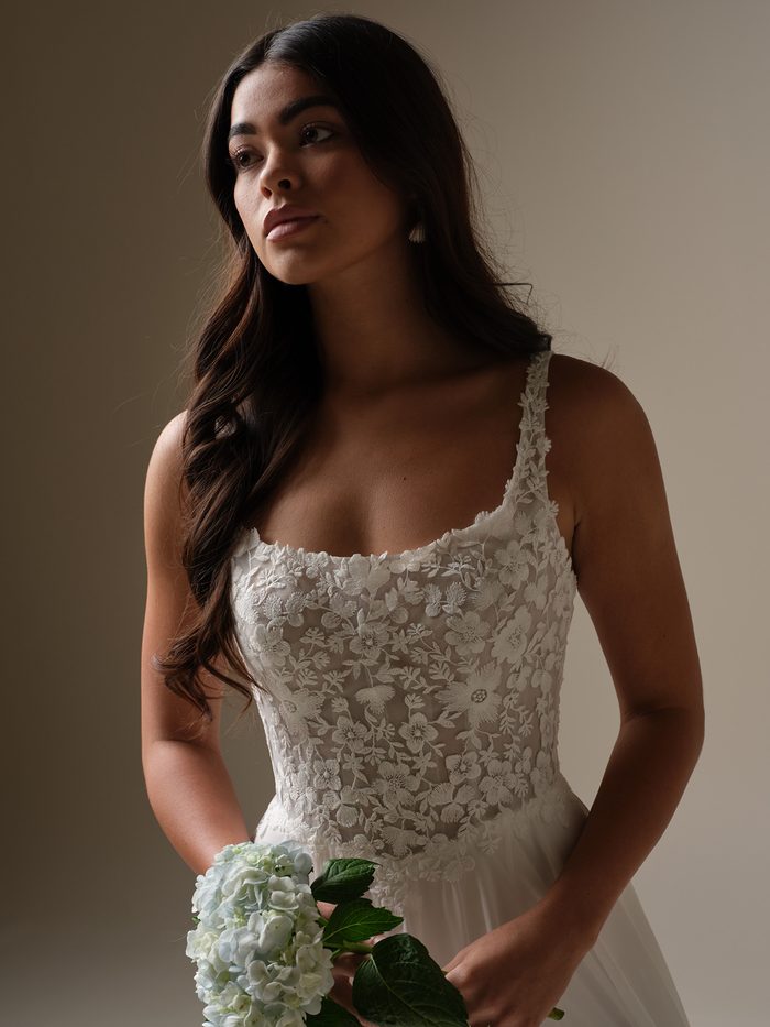 Close-up of a bride holding a hydrangea bouquet, showing the floral lace bodice and square neckline of the chiffon Cory wedding dress by Rebecca Ingram.
