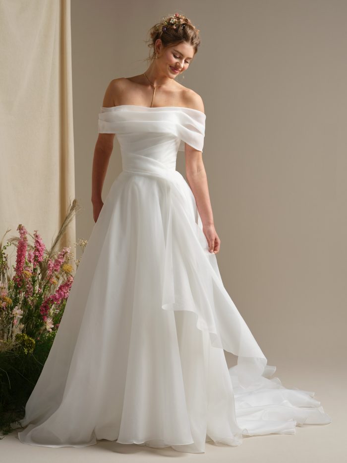 Dezi by Rebecca Ingram ballgown wedding dress with an off-the-shoulder neckline, ruched organza bodice, flowing A-line skirt, hidden pockets, and a romantic cathedral-length train.