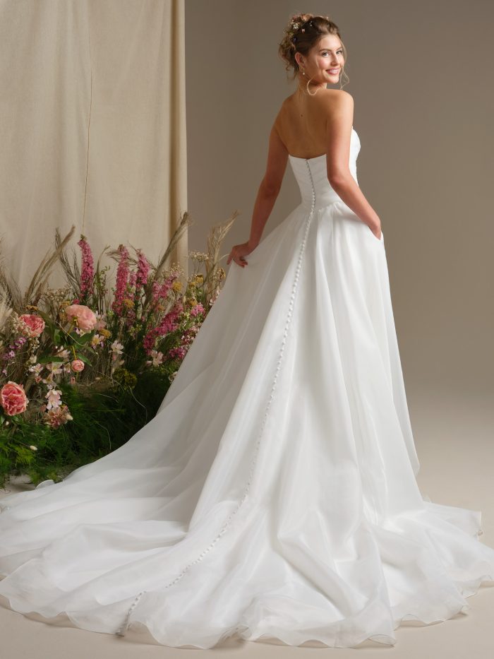 Dezi by Rebecca Ingram organza ballgown wedding dress featuring an off-the-shoulder design, covered button back detail, voluminous skirt, hidden pockets, and an extended cathedral train.