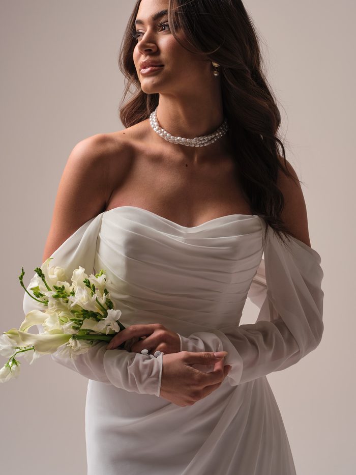 Close-up of a bride wearing a pearl choker with the draped chiffon bodice and detachable off-the-shoulder sleeves of the Francesca wedding dress by Rebecca Ingram, holding a white floral bouquet.