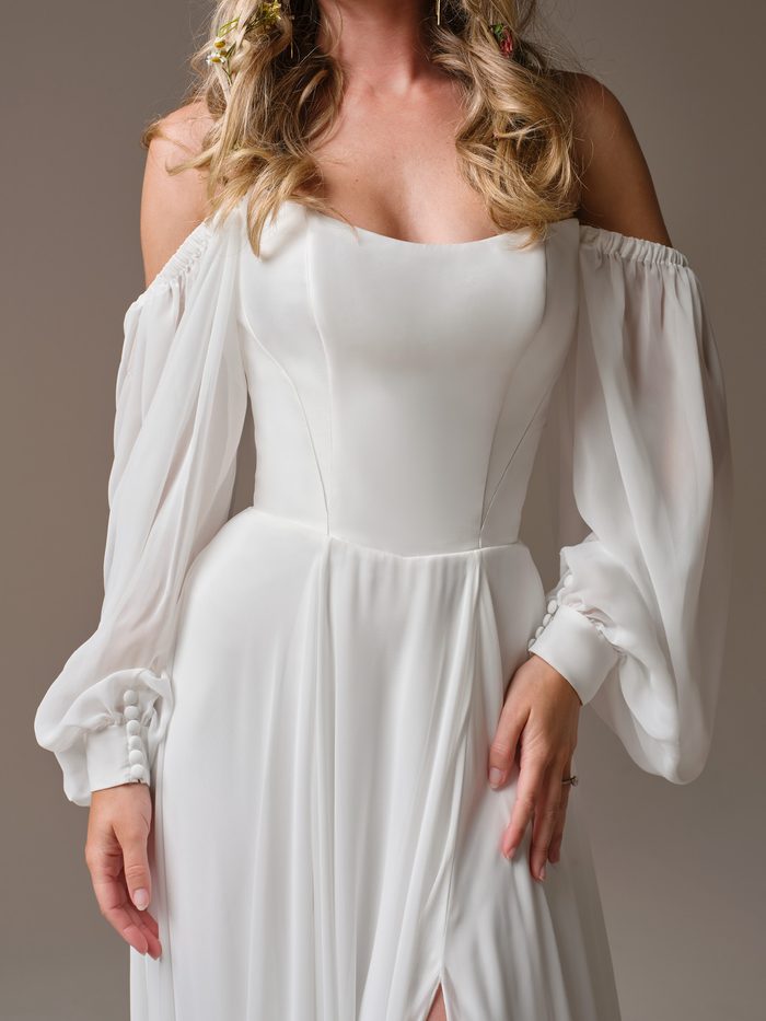 Bride wearing the Georgina A-line wedding dress by Rebecca Ingram, a chiffon gown with a straight neckline, leg slit, and flowing train, styled with wildflowers in her hair.