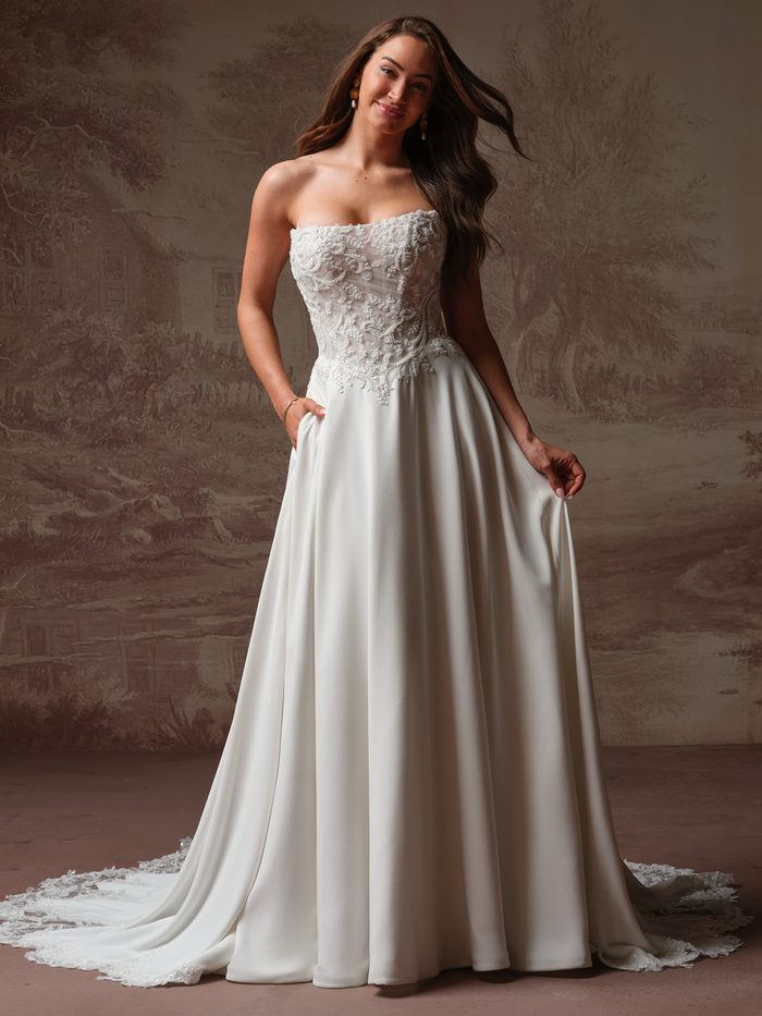 Front view of a model wearing the Hazel A-line Wedding Dress by Rebecca Ingram in ivory, featuring a strapless beaded lace bodice with sweetheart neckline and a smooth crepe A-line skirt with hand pockets and a lace-trimmed hem train.