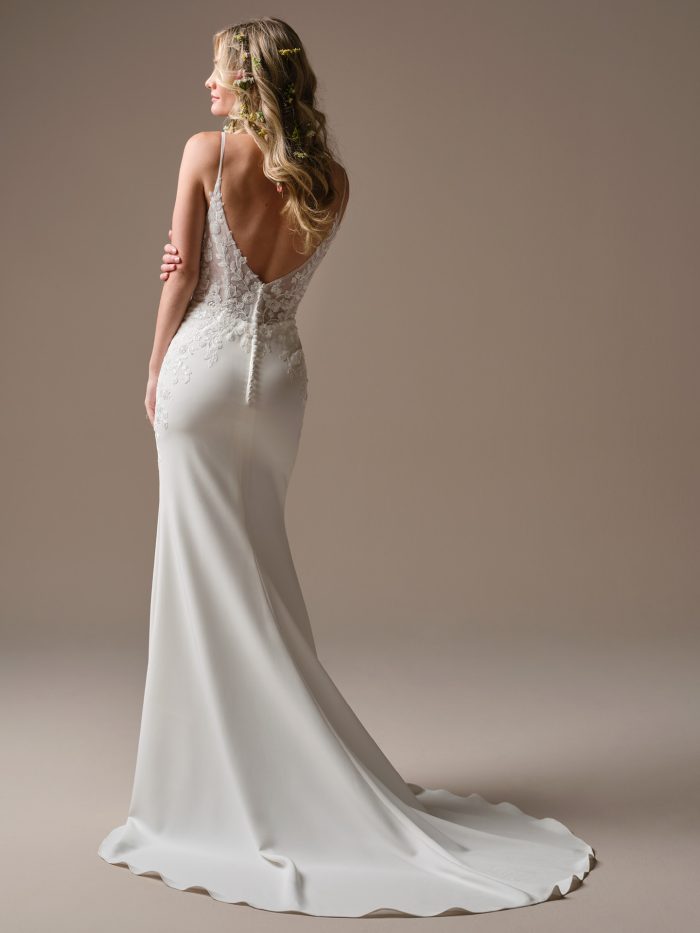 Holly by Rebecca Ingram crepe sheath wedding dress with a low V-back, floral lace appliqués, spaghetti straps, covered button detailing, and sweeping train.
