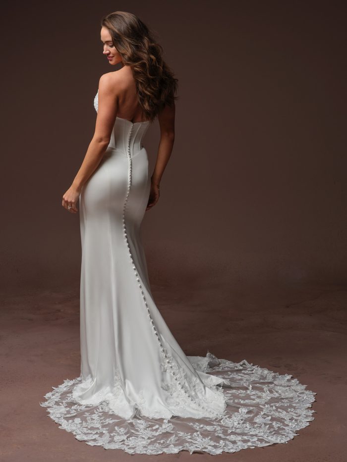 Back view of bride wearing the Isolde strapless crepe sheath wedding dress by Rebecca Ingram, featuring a sweetheart back neckline, covered button closure, and an illusion lace train.