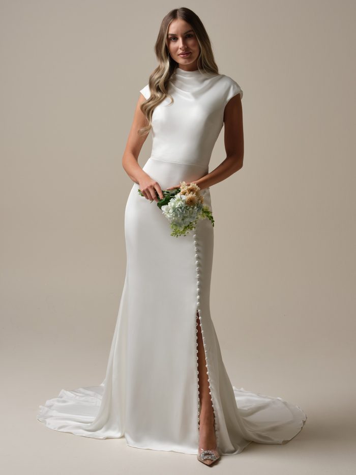 Jett Leigh by Rebecca Ingram sheath wedding dress featuring a high neckline, minimalist crepe fabric, cap sleeves, side slit with covered button detail, fitted silhouette, and elegant train.