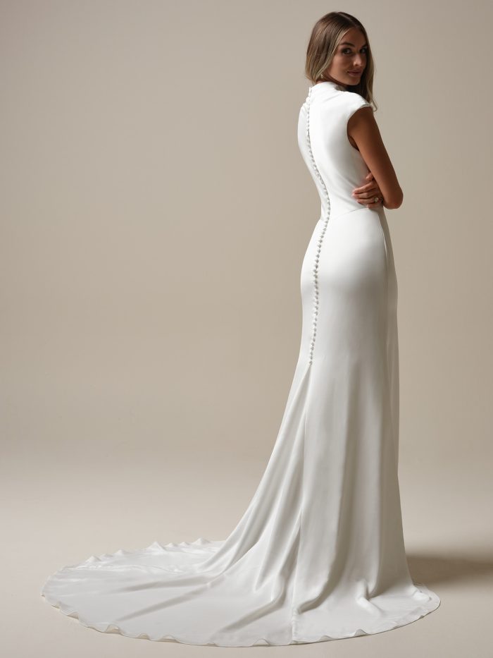 Jett Leigh by Rebecca Ingram crepe sheath wedding dress with a high neckline, cap sleeves, covered buttons down the back, figure-flattering silhouette, and sweeping train.