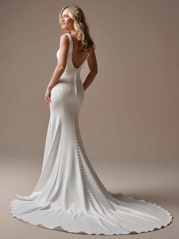 Lucille by Rebecca Ingram crepe sheath wedding dress with a low scoop back, wide straps, covered button detailing, figure-flattering silhouette, and elegant train.