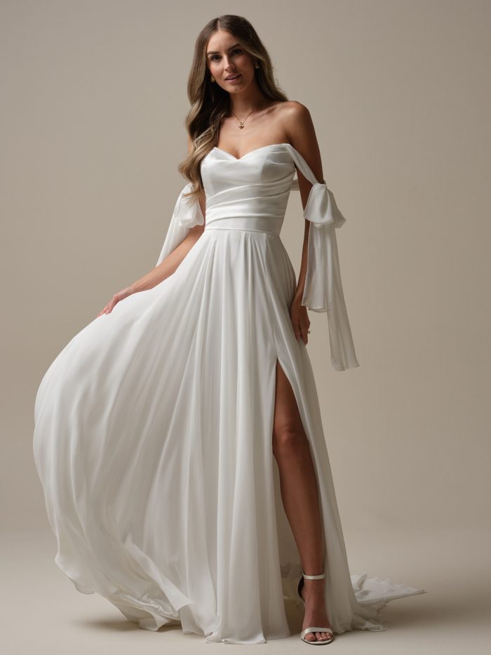Melody by Rebecca Ingram A-line wedding dress featuring a sweetheart neckline, detachable draped off-the-shoulder sleeves, chiffon skirt with a thigh-high slit, and flowing train.