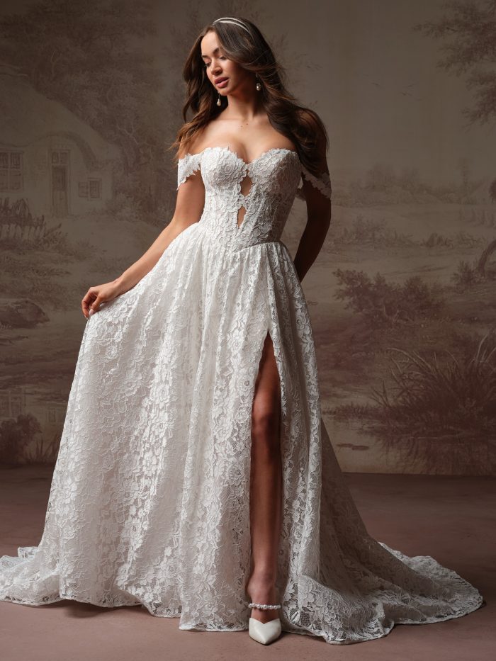Full-length view of a bride wearing the Narelle wedding dress by Rebecca Ingram, an A-line lace gown with an off-the-shoulder sweetheart neckline, keyhole bodice, and a leg slit skirt with train.