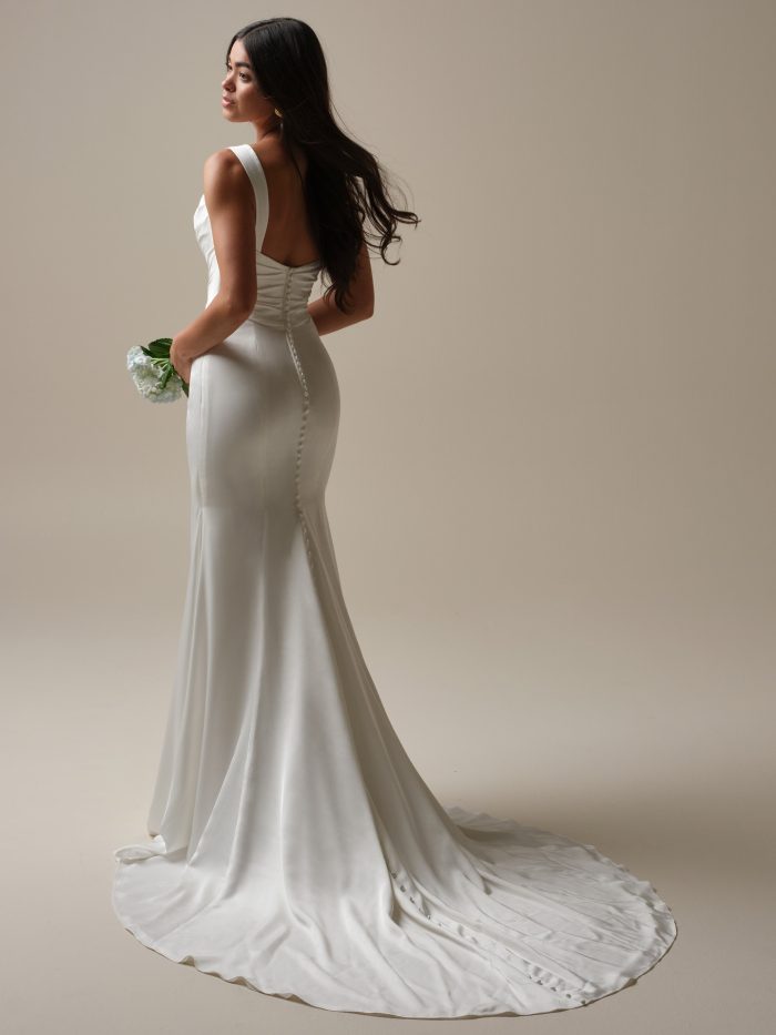 Tommie by Rebecca Ingram satin sheath wedding dress with a low scoop back, covered button detailing, wide straps, figure-flattering silhouette, and flowing train.