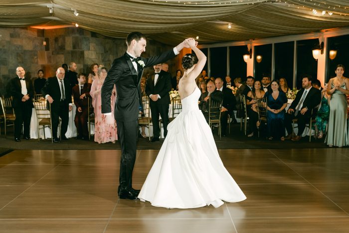 Wedding photo of a groom twirling his bride during their first dance in a strapless satin ballgown wedding dress by Maggie Sottero, surrounded by guests at the reception.