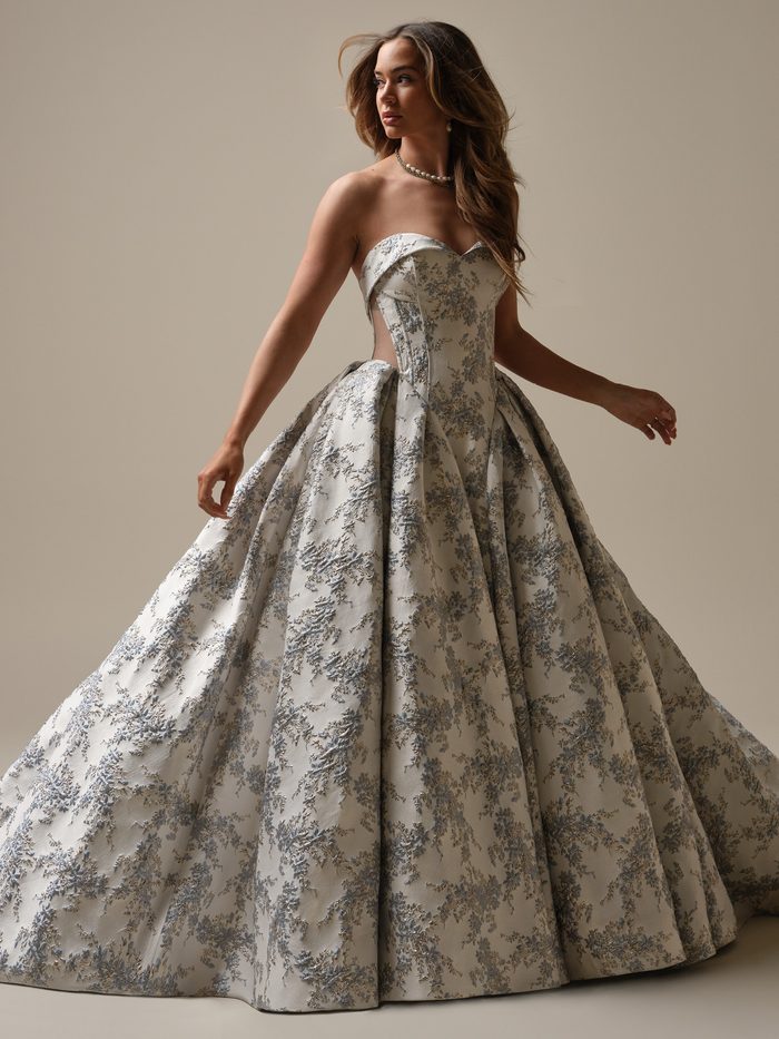 Front view of a model wearing the Aberdeen Ballgown Wedding Dress by Sottero and Midgley in blue, featuring a metallic floral brocade fabric with silver and gold accents, a strapless folded-cuff bodice with sheer side cutouts, and a dramatically full skirt.
