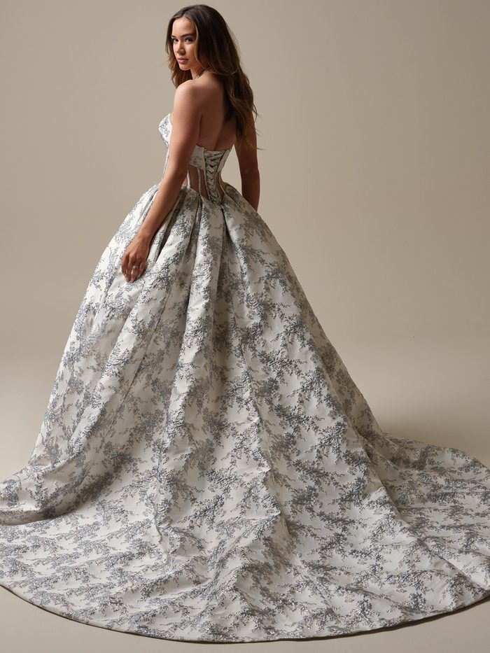 Back view of a model wearing the Aberdeen Ballgown Wedding Dress by Sottero and Midgley in blue, showing the strapless metallic brocade bodice with a corset-style lace-up back and sheer side cutouts, paired with a voluminous floral jacquard ball skirt with a train.