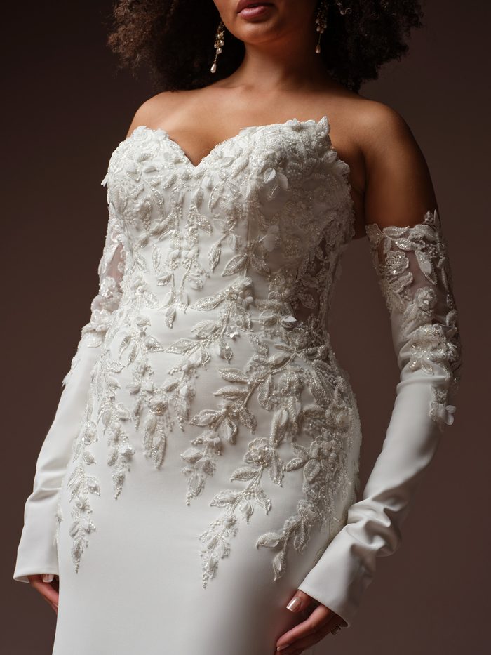 Close-up of a bride wearing the Blair sheath wedding dress by Sottero and Midgley, showing detailed beaded floral lace on the bodice and off-the-shoulder sleeves.