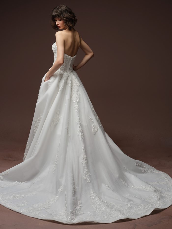 Back view of bride wearing the Gwyneth strapless ballgown wedding dress by Sottero and Midgley, featuring a beaded lace bodice, sweetheart back neckline, covered button closure, pockets, and an embellished lace train.