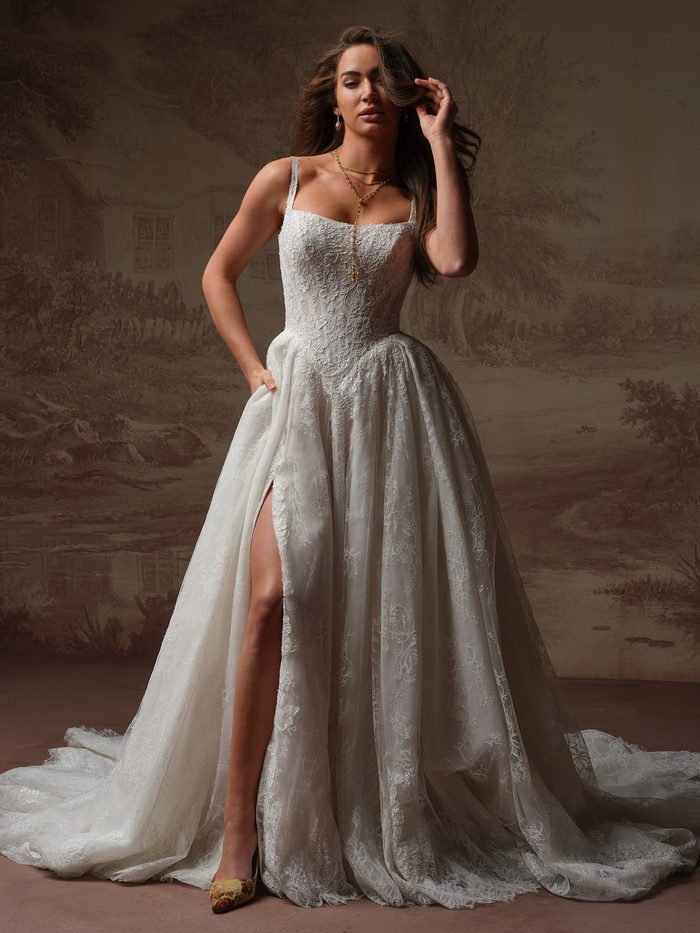 Model wearing the Ivandra ballgown wedding dress by Sottero and Midgley, featuring a straight-across lace bodice with thin straps and a full lace ballgown skirt with front slit and train.