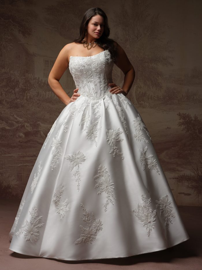 Front view of a curve model wearing the Lucinde ballgown wedding dress by Sottero and Midgley, featuring a strapless beaded bodice and a full satin skirt scattered with sparkling snowflake-like lace appliqués.