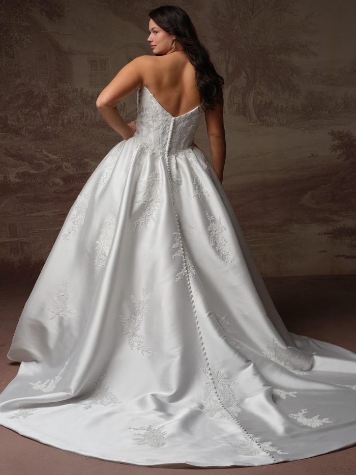 Back view of a curve model wearing the Lucinde ballgown wedding dress by Sottero and Midgley, showing the fitted beaded bodice with covered button closure and a dramatic satin train adorned with scattered lace appliqués.