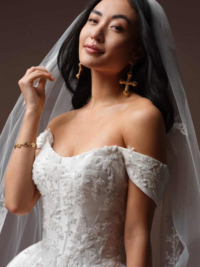 Bride wearing Lucinde by Sottero and Midgley, an off-the-shoulder wedding dress with beaded floral lace bodice, cap sleeves, and matching lace-trimmed veil.