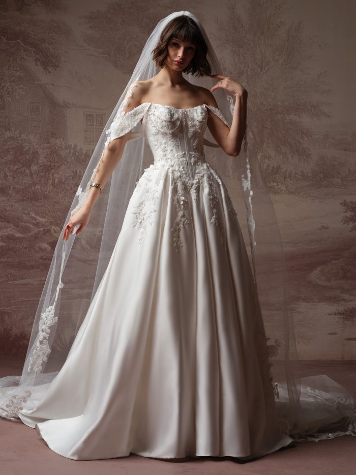 Full-length view of a bride wearing the Petra wedding dress by Sottero and Midgley, featuring an off-the-shoulder beaded lace bodice, satin ball gown skirt, and a lace-edged cathedral veil.