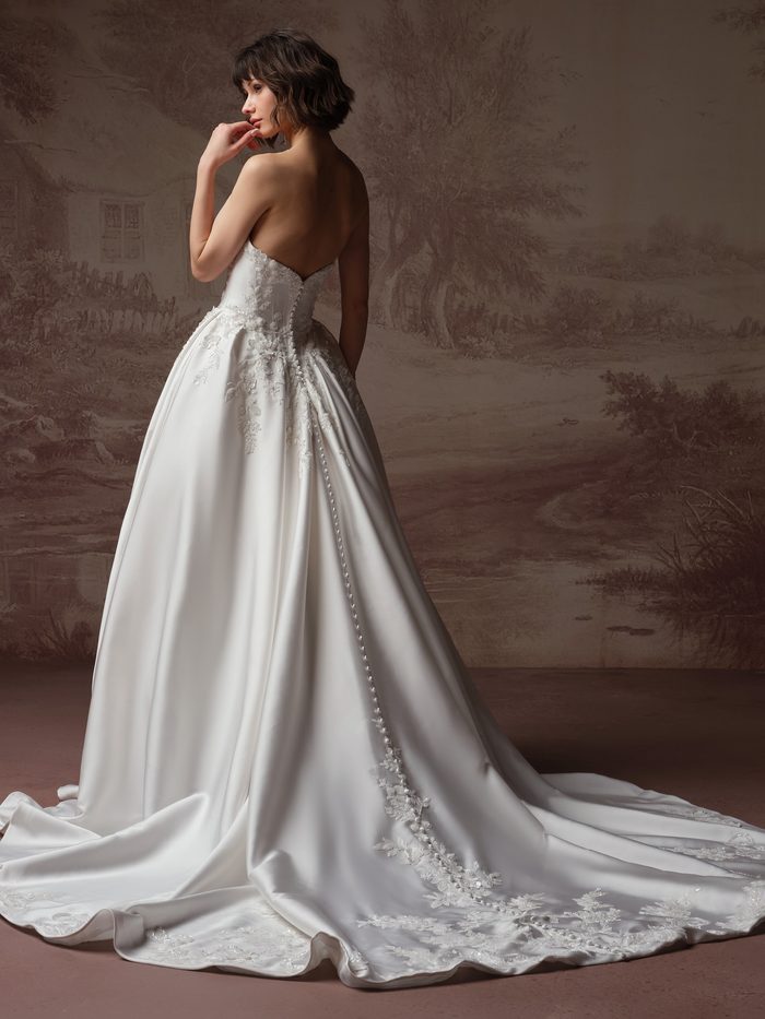 Back view of a model wearing the Petra Ballgown Wedding Dress by Sottero and Midgley in ivory, showing the strapless lace bodice with a row of covered buttons and lace trim down the back seam, and a voluminous satin ball skirt with a long train.