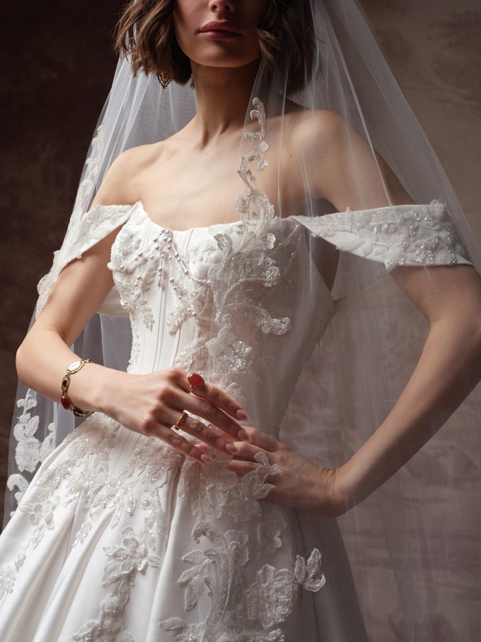 Close-up of a bride wearing the Petra wedding dress by Sottero and Midgley, highlighting the beaded lace bodice, off-the-shoulder sleeves, and floral lace-trimmed veil.