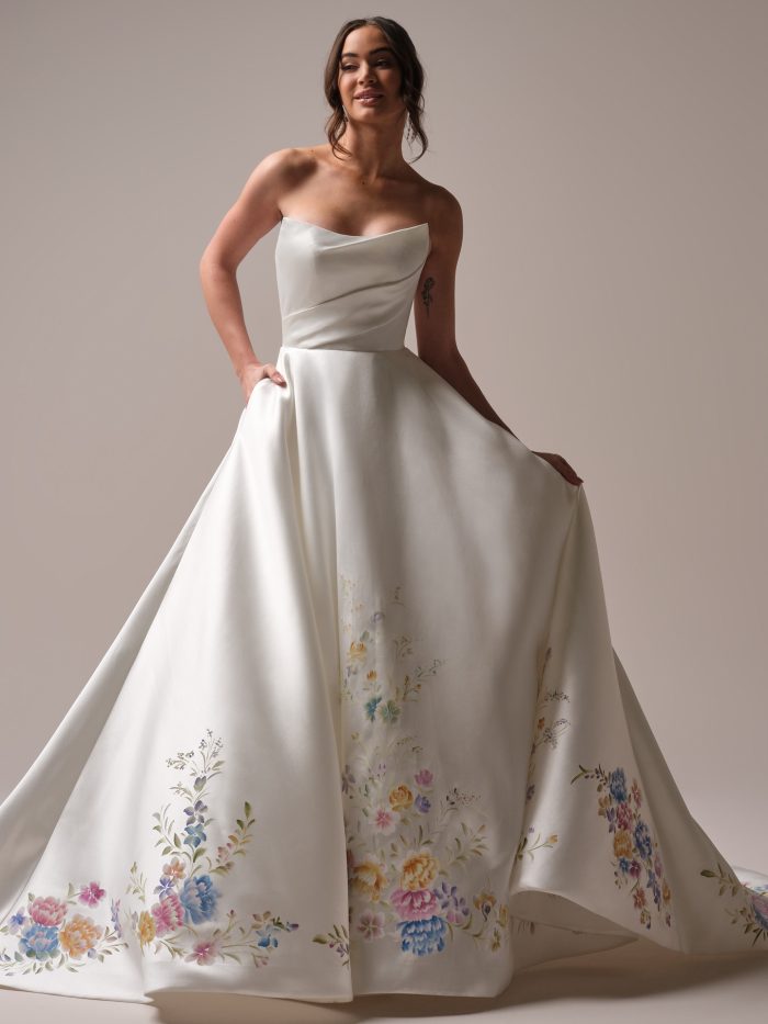 Front view of a bride wearing the Radiance A-line wedding dress by Sottero and Midgley, featuring a strapless satin bodice with a draped cowl neckline and a full skirt printed with a colorful watercolor floral design at the hem and train.