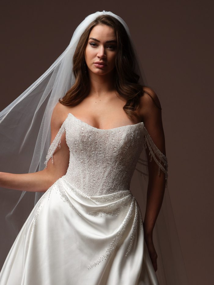 Bride wearing Rumi by Sottero and Midgley, a beaded wedding dress with draped crystal fringe cap sleeves, satin draped skirt, and veil.