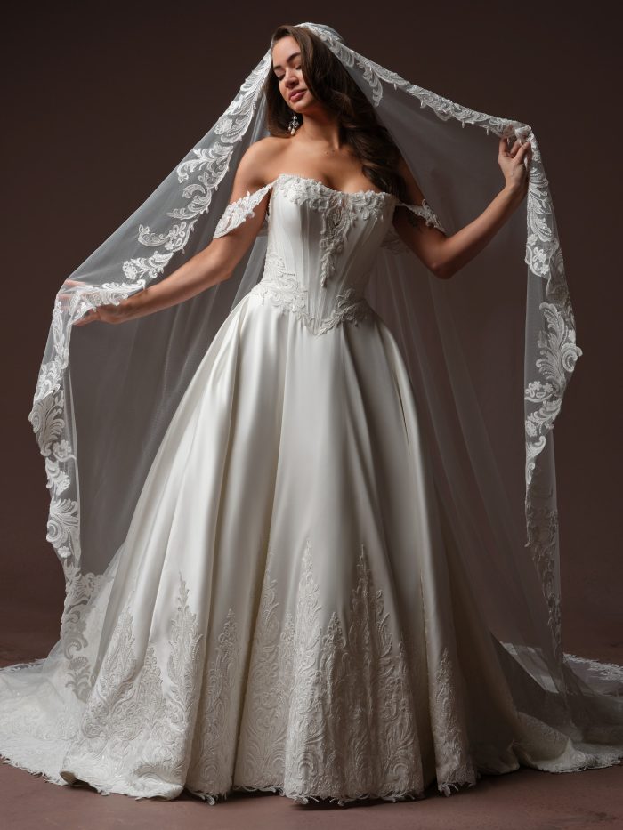 Front view of a bride wearing the Sandrine ballgown wedding dress by Sottero and Midgley, featuring an off-the-shoulder beaded lace bodice, a full satin skirt with lace hem detailing, and a matching lace-edged cathedral veil.