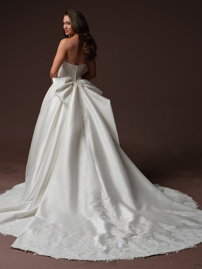 Back view of bride wearing the Sandrine strapless satin ballgown by Sottero and Midgley with a detachable train, featuring a lace bodice, sweetheart back neckline, covered button closure, and an oversized bow with cathedral-length embellished lace hem.