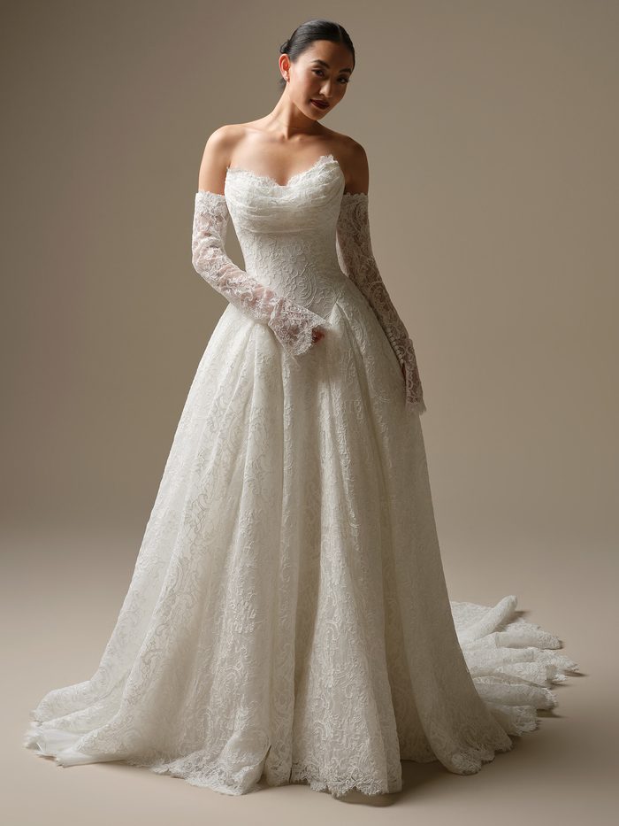 Front view of a model wearing the Tamar A-line Wedding Dress by Sottero and Midgley in ivory, featuring an off-the-shoulder scalloped lace bodice with sweetheart neckline, detached sheer lace long sleeves with scalloped cuffs, and a full lace A-line skirt with scalloped hem and train