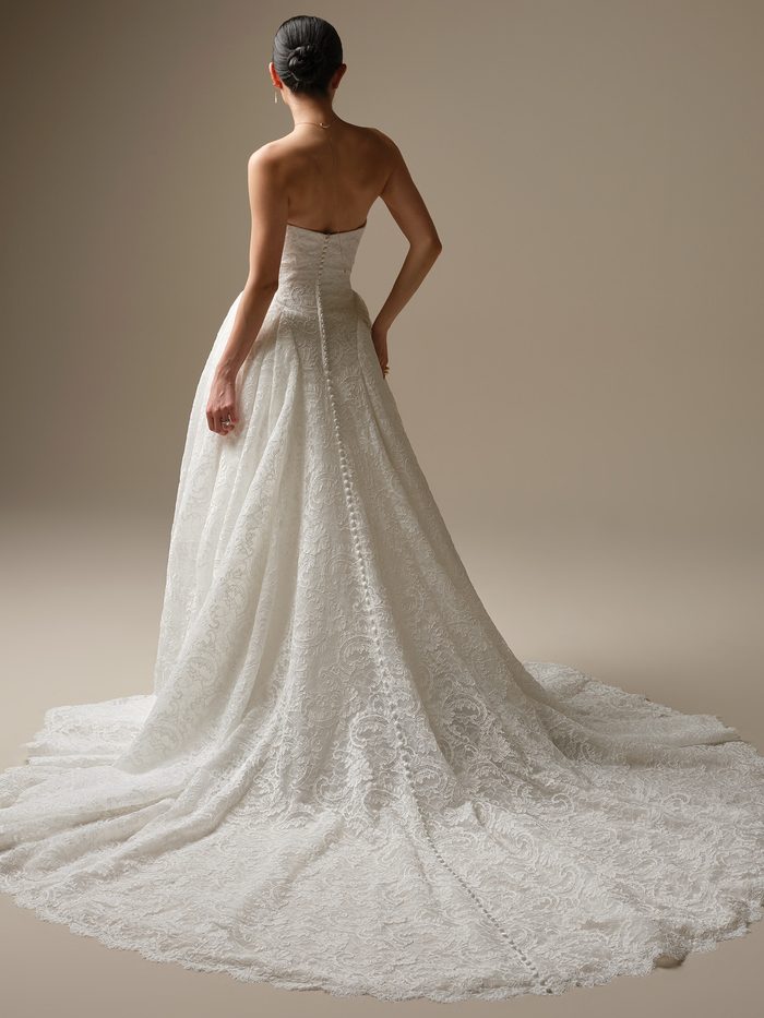 Back view of a model wearing the Tamar A-line Wedding Dress by Sottero and Midgley in ivory, showing the strapless lace bodice with a row of covered buttons down the back, and a flowing lace A-line skirt with scalloped hem and long train.