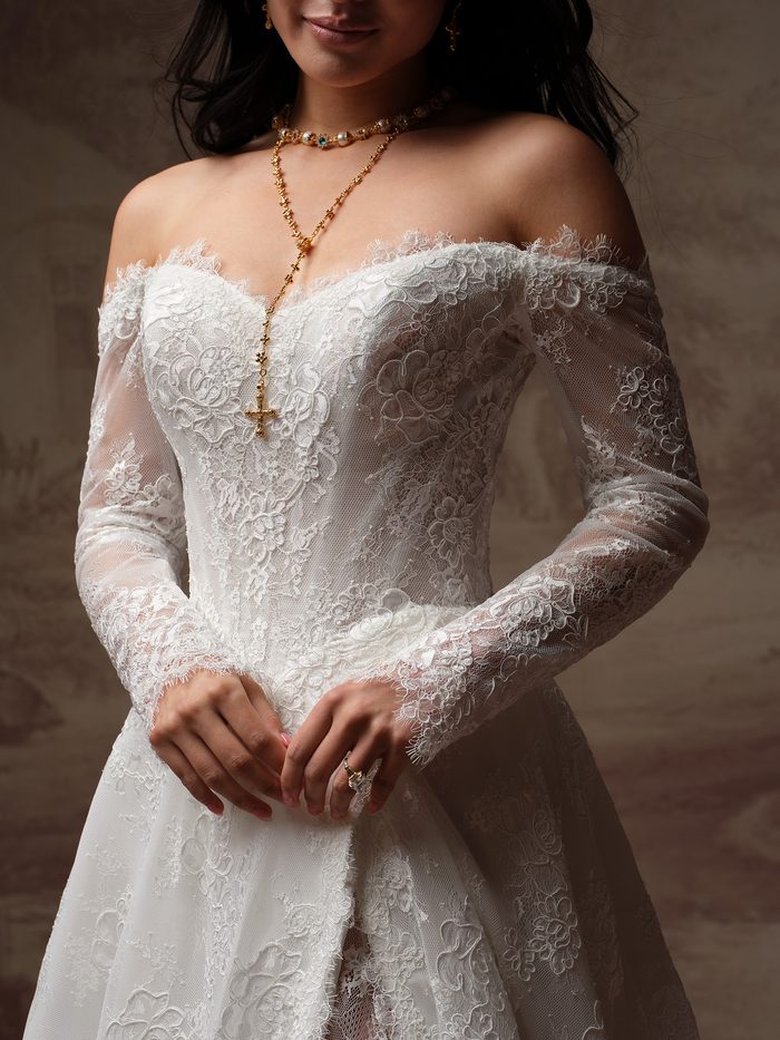 Close-up of the Vespera A-line wedding dress by Sottero and Midgley, showing the off-the-shoulder lace neckline, sheer long sleeves, and floral lace appliqué on the bodice.