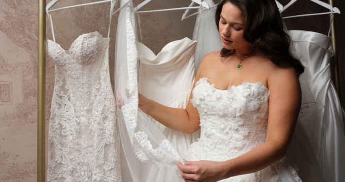 A bride in a strapless, lace-appliqué wedding gown (Benton by Maggie Sottero) examines a lace veil while browsing a rack of other wedding dresses in a bridal boutique.