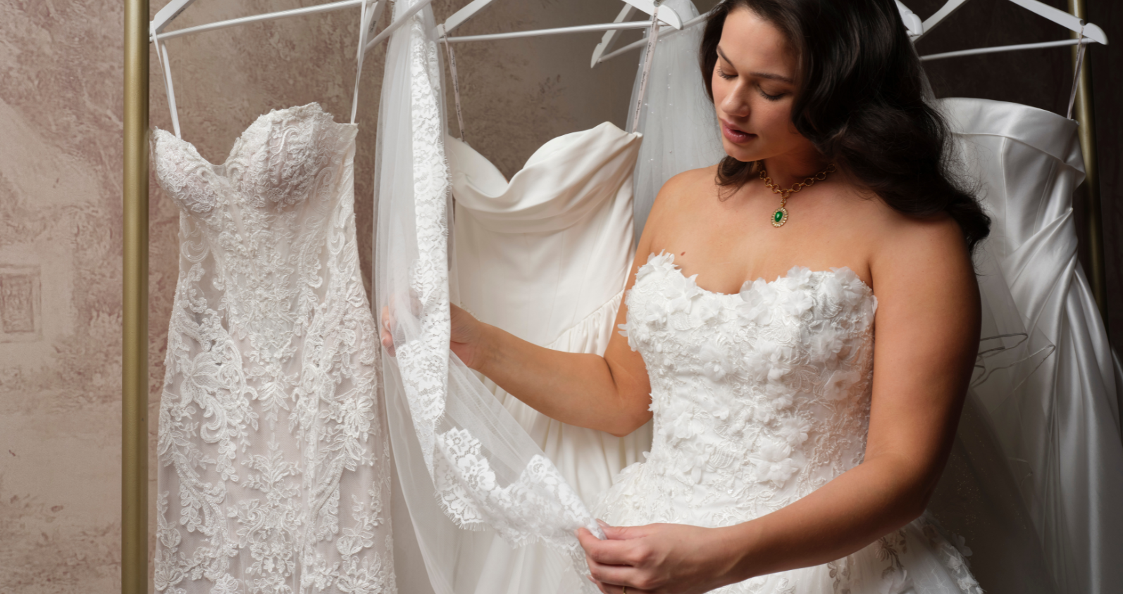 A bride in a strapless, lace-appliqué wedding gown (Benton by Maggie Sottero) examines a lace veil while browsing a rack of other wedding dresses in a bridal boutique.