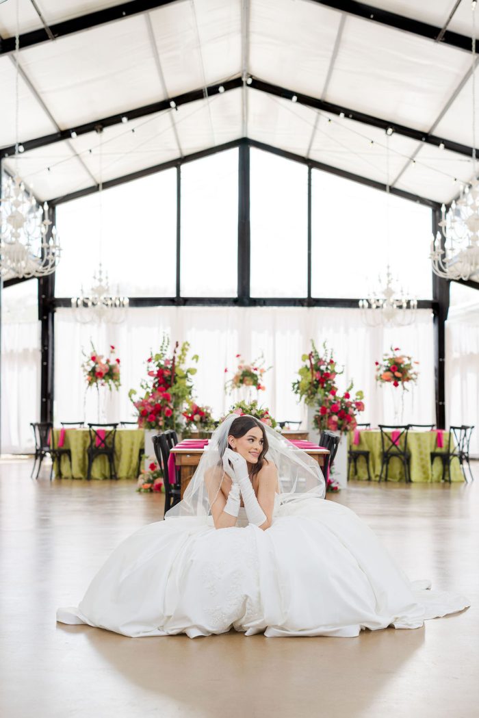 Bride in a Maggie Sottero ball gown wedding dress with opera-length gloves and veil, seated on the floor of a glass-walled reception tent decorated with vibrant pink and green florals, photographed by Winterlyn Photography for Chicago Style Weddings.