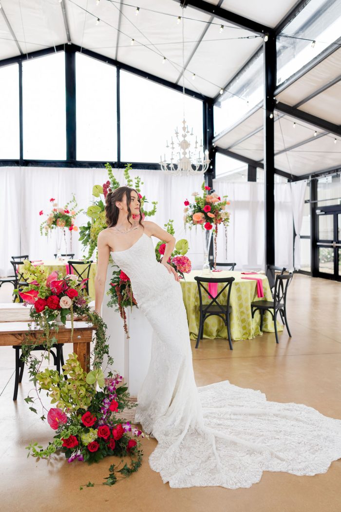 Bride in a fitted lace one-shoulder Maggie Sottero wedding gown with a long train, posing beside a floral-draped sweetheart table in a glass-walled tent reception space, photographed by Winterlyn Photography for Chicago Style Weddings.