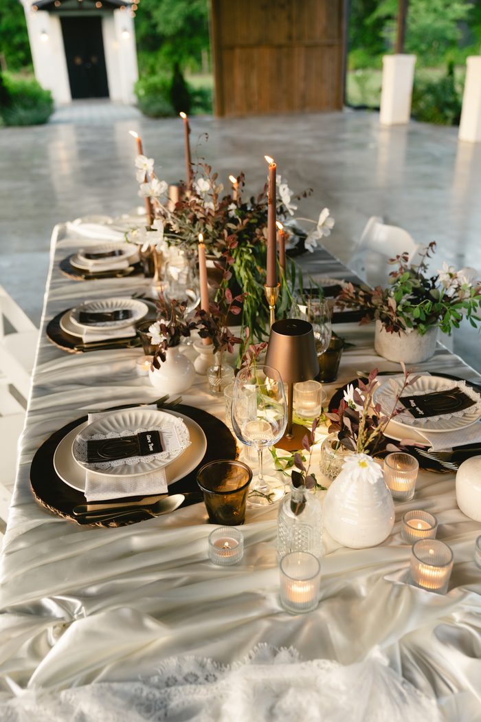 Elegant wedding reception table setting with black chargers, white scalloped plates, dusty rose taper candles, greenery and white floral centerpieces, and votive candles along a draped white tablecloth.