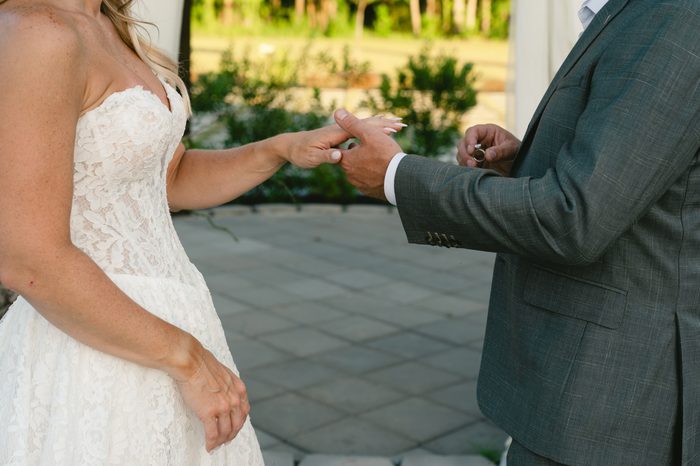 Close-up of a groom placing a wedding ring on the bride's finger, the bride wearing a lace Narelle by Rebecca Ingram gown.