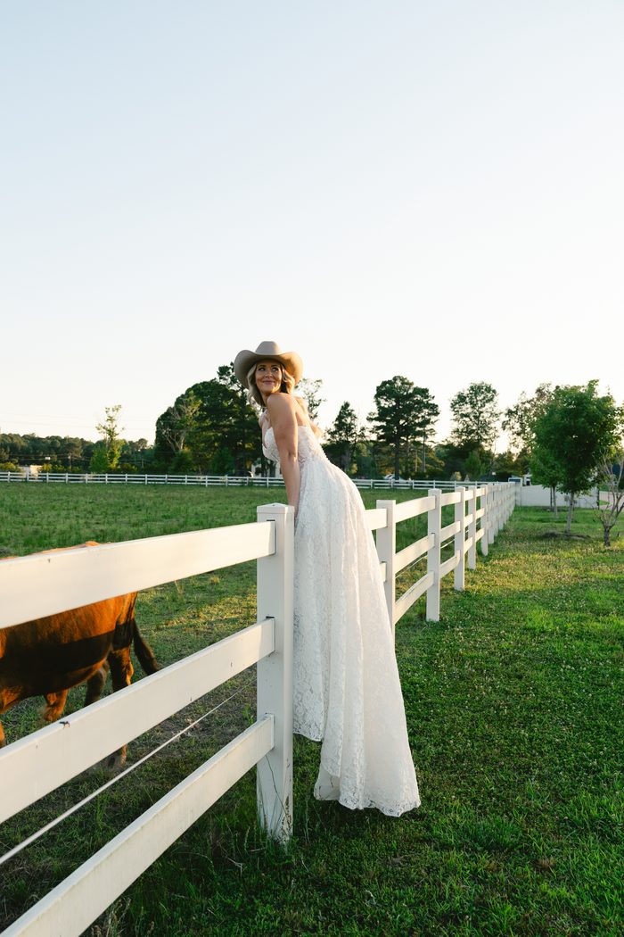 Bride in a lace wedding gown Narelle by Maggie Sottero and cowboy hat leans against a white fence post, smiling back over her shoulder as a horse grazes nearby in a green pasture.