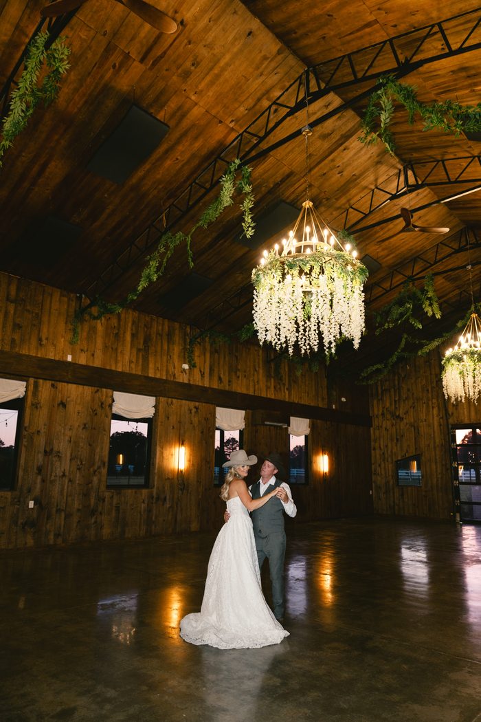 Bride and groom in cowboy hats share their first dance on a wooden barn dance floor, lit by a chandelier draped in white wisteria, the bride wearing a lace Narelle by Maggie Sottero gown.