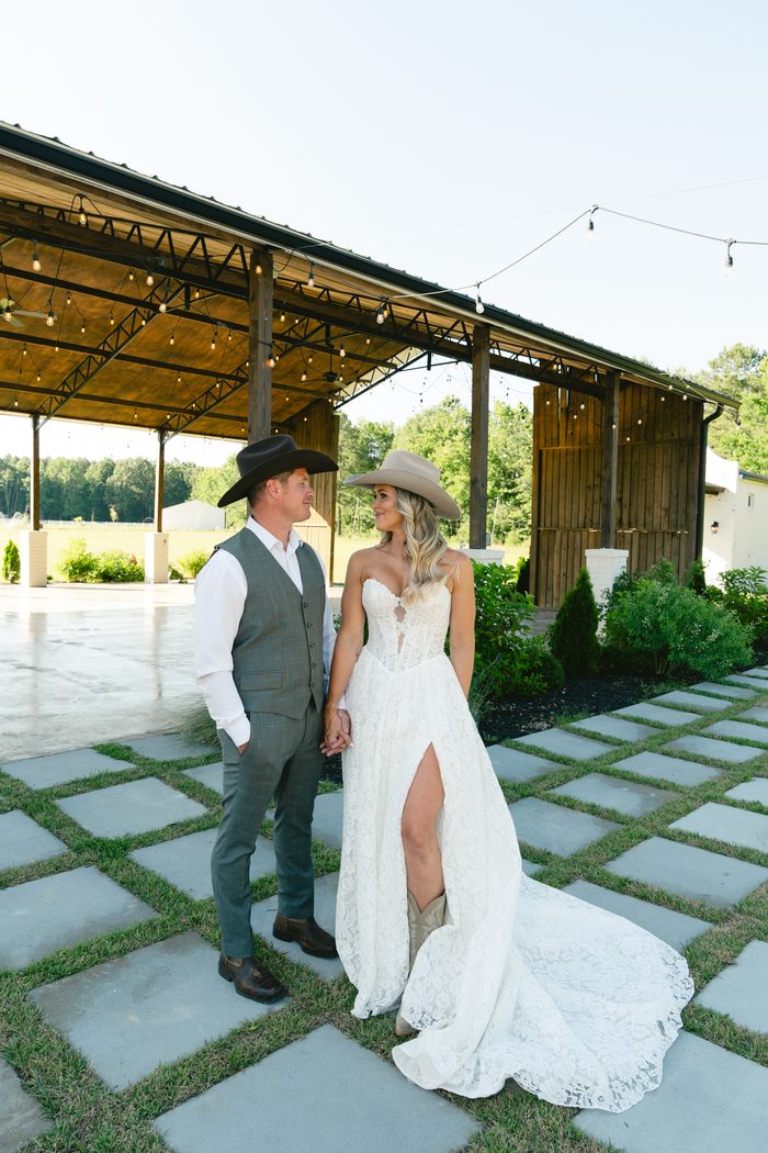 Bride and groom in cowboy hats hold hands on a checkered stone pathway outside a string-lit barn venue, the bride wearing a lace Narelle by Rebecca Ingram gown with a leg slit and cowboy boots.