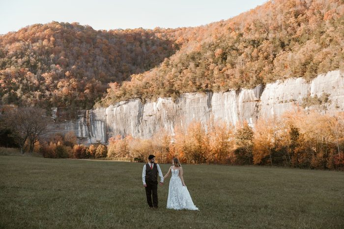 Wide outdoor portrait of a couple walking hand in hand across a grassy field beneath towering limestone cliffs and vibrant autumn foliage. One person wears the Natalie by Maggie Sottero wedding dress, featuring a fitted bodice and floral lace A-line skirt, set against a scenic fall landscape with golden and orange leaves.