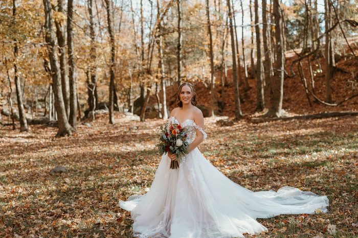 Bride standing in a sunlit autumn woodland wearing the Wicklow wedding dress by Maggie Sottero, an off-the-shoulder floral lace ballgown with a full tulle skirt and sweeping train. The bride holds a bouquet of rust, orange, ivory, and greenery blooms, surrounded by fallen leaves and golden fall foliage.
