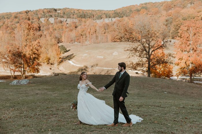 Bride wearing the Wicklow wedding dress by Maggie Sottero walks hand in hand with a partner across a grassy hillside, showcasing the gown’s off-the-shoulder floral lace bodice, full tulle skirt, and flowing train. Vibrant autumn trees and rolling countryside create a scenic fall wedding backdrop.